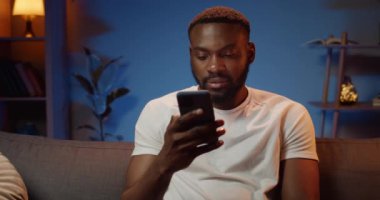 Kanepede oturan ve telefon kumandasını kullanan yakışıklı adamın ön görüntüsü. Sakallı Afrikalı adam evde vakit geçirirken arkadaşlarına sesli mesaj bırakıyor. Teknoloji kavramı.