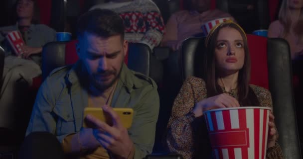 Hombre viendo deporte partido durante la fecha con su novia en el cine. Chico usando smartphone y chica comiendo palomitas de maíz mientras ve películas en el cine. Concepto de ocio y entretenimiento . — Vídeo de stock