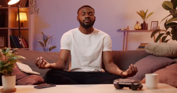 Mladý pohledný muž cvičí dýchání, zatímco sedí doma na pohovce. Vousatý Afričan dělá jógu, zatímco relaxuje ve volném čase. Koncepce zdravého životního stylu a volného času. — Stock video