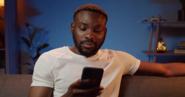 Μπροστά όψη του όμορφου Αφρικανού άνδρα που κοιτάζει την οθόνη του smartphone ενώ κάθεται στον καναπέ. Γενειοφόρος νεαρός συνομιλούν στα κοινωνικά δίκτυα, ενώ περνούν ελεύθερο χρόνο στο σπίτι. — Αρχείο Βίντεο