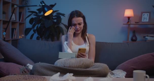 Vista frontal de la joven mujer molesta usando pañuelos de papel mientras sostiene el teléfono inteligente. Niña infeliz llorando y teniendo crisis emocional después de discutir con el amante mientras está sentado en el sofá . — Vídeo de stock