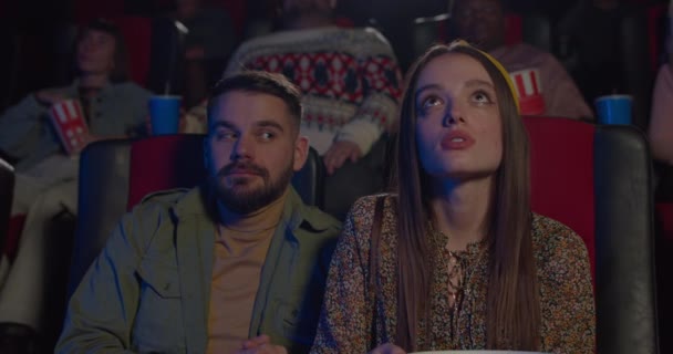 Un tipo barbudo abrazando a una chica guapa sentada a su lado en el cine. Chica joven que se ve feliz en la cita en el cine mientras su novio abrazándola. Concepto de sentimientos humanos . — Vídeos de Stock