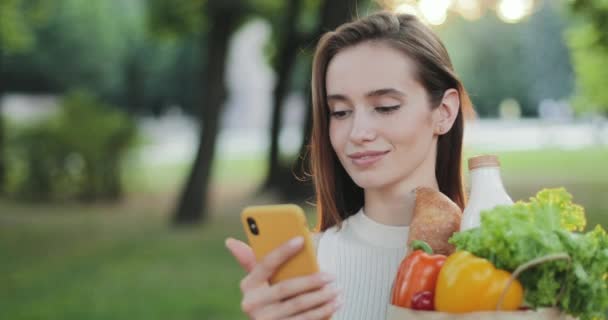 Közelkép a gyönyörű fiatal nő segítségével okostelefon és mosolygós. Elég vidám lány nézi a telefonképernyőt, miközben papírzacskót tart étellel és mosollyal. A vásárlás fogalma. — Stock videók