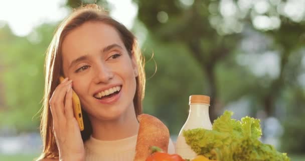 Nahaufnahme von schönen Mädchen, die am Telefon sprechen und Papiertüten mit Lebensmitteln in der Hand halten. Lächelnde junge Frau kommuniziert auf Smartphone und hält ihre Einkäufe im Freien. — Stockvideo