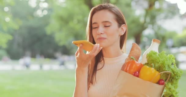 Csinos fiatal nő beszél, miközben okostelefon hangvezérét használja, és papírzacskót tart étellel. Vidám Y-generációs lány diktálja az üzenetet, miközben áll a kezében a vásárlással.. — Stock videók