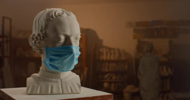 Cabeça de escultura de gesso branco de pessoa famosa vestindo máscara médica em pé sobre pequena mesa no estúdio escultor. Conservado da arte no qurantine . — Vídeo de Stock