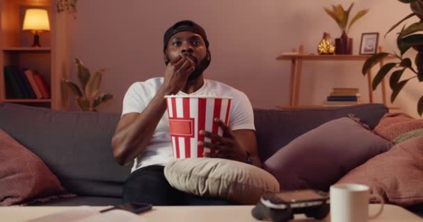 Spännande kille äter popcorn från pappersskål och ler medan du tittar på TV-skärmen. Glad skäggig man i mössa tittar på bra film och ler och ser nöjd ut hemma. — Stockvideo