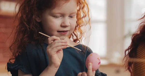 Gadis kecil berambut merah ceria dengan ibunya melukis telur Paskah di latar belakang dapur. Selamat easter Stok Gambar
