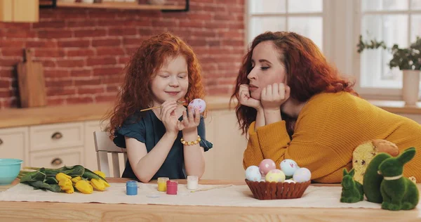 Ibu dan putrinya sedang mempersiapkan Paskah. Gadis kecil melukis telur Paskah di dapur yang nyaman. Ibu tersenyum melihat putrinya. Stok Foto Bebas Royalti