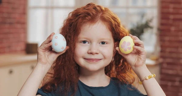 Portretul unei fetițe roșcate vesele care se joacă cu ou de Paști pe fundalul bucătăriei. Ea se bucură și se distrează la cameră. Happy Easter Fotografie de stoc