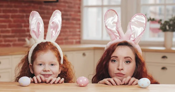Potret ibu lucu dan putrinya mengenakan telinga kelinci dengan telur Paskah melihat ke kamera. Keluarga bahagia bersenang-senang di dapur yang nyaman. Selamat easter Stok Foto Bebas Royalti