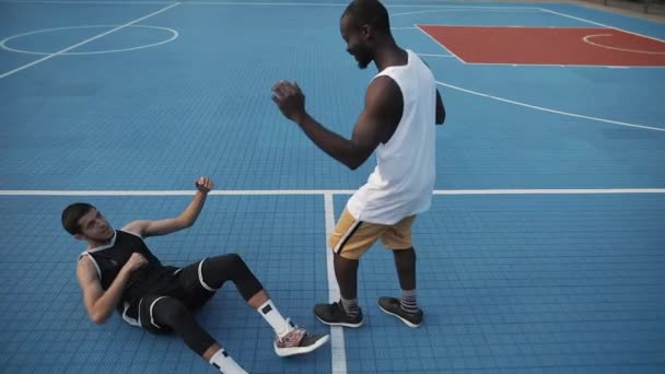 Schöner muskulöser Afroamerikaner gibt Hand und hilft dem jungen Mann aus dem Kaukasus beim Street Sports Basketball Court auf die Beine. Gesunder Lebensstil und Sportkonzept. Zeitlupe. — Stockvideo