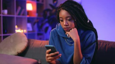 Evde vakit geçirirken akıllı telefon kullanan Afro-Amerikan kız portresi. Genç ve çekici bir kadın kanepesinde otururken ekrana bakıyor ve internette geziniyor. Yakınlaştır.