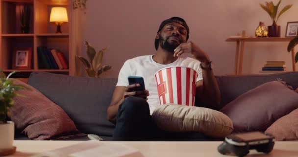 Vousatý mladý muž zívá a třese hlavou, zatímco sedí na pohovce s popcorn mísa před televizí.Nudit africký chlap pomocí svého smartphonu při sledování nezajímavého filmu . — Stock video