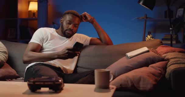 사려깊은 젊은이가 소파에 앉아 있는 동안 스마트폰을 사용하는 모습. 수염난 아프리카 남자 가 집에서 자유 시간을 보내면서 핸드폰 화면을 스크롤하고 있어요. 여가 와 기술에 대한 개념. — 비디오