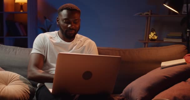 Frontansicht eines gutaussehenden Kerls, der spät abends mit seinem Laptop im Netz surft. Junger afrikanischer Mann sitzt auf dem Sofa, schaut auf den PC-Bildschirm und tippt zu Hause. Freizeitkonzept. — Stockvideo