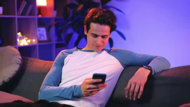 Portrait of millennial caucasian man using his modern smartphone while spending time at home. Handsome guy looking at phone screen and browsing internet while sitting on sofa. Zoom in. — Stock Video