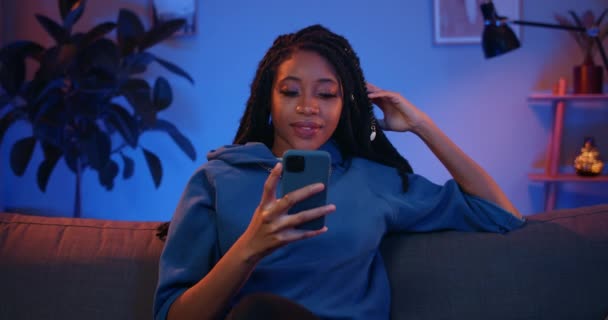 Mujer africana joven desplazándose noticias mientras usa su teléfono inteligente moderno. Chica milenaria con rastas mirando a la pantalla y sonriendo mientras está sentado en el sofá. Fondo de origen . — Vídeos de Stock