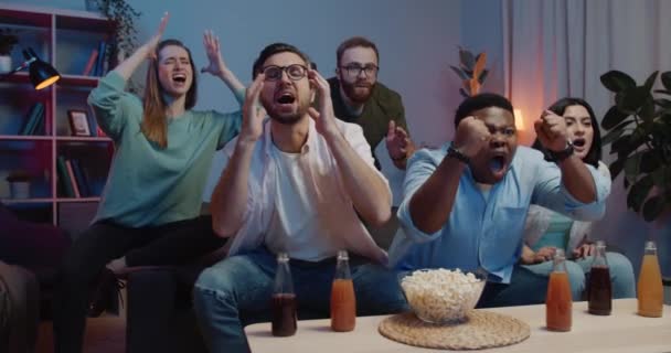 Gruppo multietnico di uomini e donne che sostengono la squadra mentre guardano lo sport in TV. Eccitato gruppo di amici seduti sul divano davanti al tavolo con popcorn e bevande e arrabbiarsi . — Video Stock