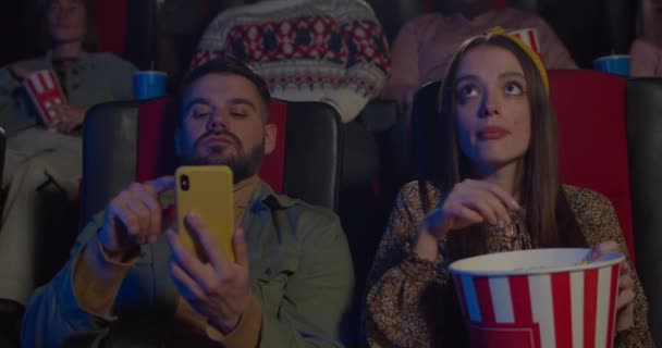 Chica comiendo palomitas de maíz y hombre usando el teléfono durante la película en el cine. Mujer joven mirando a su novio usando un teléfono inteligente y encogiéndose de hombros mientras ve la película. Concepto de ocio . — Vídeos de Stock