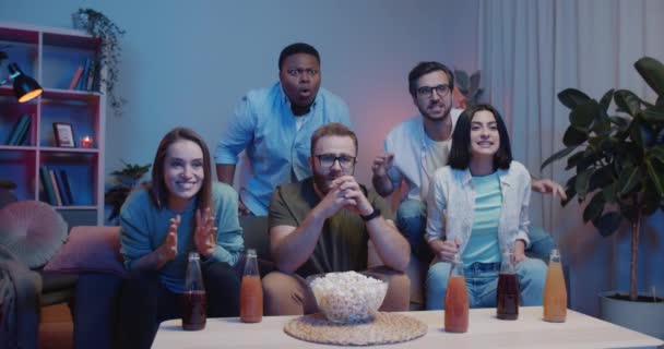 Amigos felices celebrando éxitos cerca del tipo molesto apoyando a otro equipo. Grupo de personas multiétnicas sentadas en el sofá y viendo el campeonato deportivo en la televisión. Concepto de ocio . — Vídeos de Stock