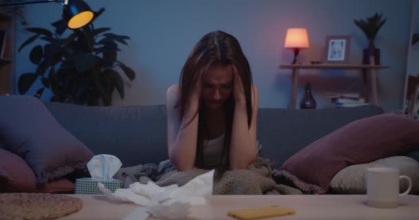 Front view of girl suffering from headache while sitting on couch at home. Young woman holding her head with hands and worrying about something.Concept of: despair, broken love, drama. — Stock Video