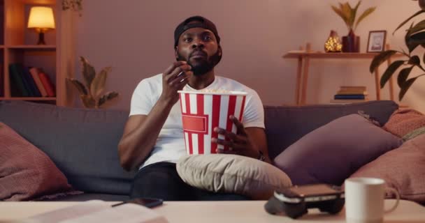 Ung afrikansk kille som ser besviken ut medan han tittar på film hemma. Snygg man i mössa äter popcorn och visar känslor medan han sitter på soffan framför TV. Begreppet fritid. — Stockvideo