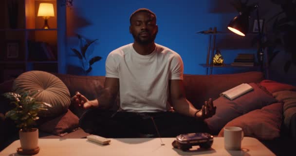 Fešák v tričku sedí na pohovce se zkříženýma nohama a cvičí jógu. Klidný afro Američan relaxuje po náročném dnu a medituje doma. Pojetí volného času. — Stock video