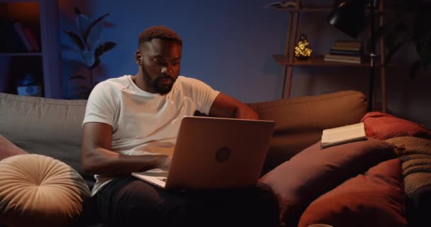 Joven barbudo usando su portátil y escribiendo mientras está sentado en el sofá en casa. Hombre africano guapo buscando concentrado mientras trabaja desde casa por la noche. Concepto de freelance . — Vídeo de stock