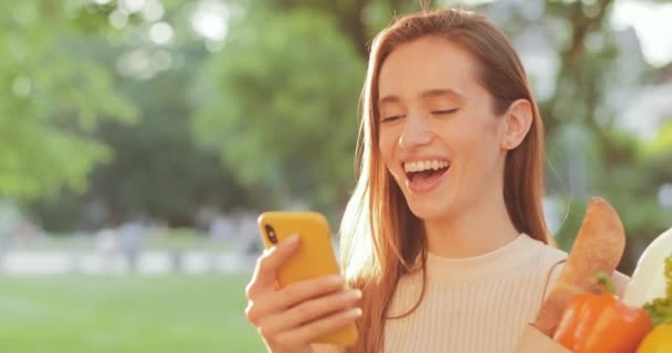 Gelukkige vrouw die grappige video kijkt of een bericht leest en lacht terwijl ze naar het scherm kijkt. mooi millennial meisje houden voedsel in zak en glimlachen terwijl staan en met behulp van haar telefoon. — Stockvideo