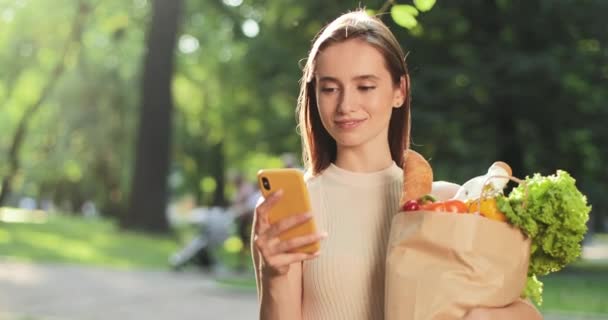 Affascinante giovane ragazza in possesso di sacchetto di carta con generi alimentari e utilizzando smartphone mentre in piedi nel parco. Ritratto di donna bruna che guarda lo schermo e naviga in rete mentre sorride alla telecamera . — Video Stock