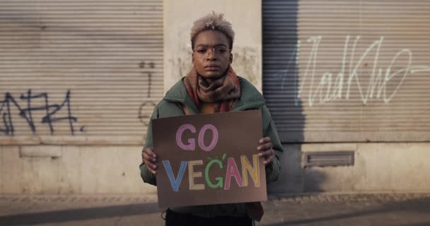 Lviv, Ucrânia - 27 de novembro de 2019: Jovem menina africana segurando Go Vegan banner sinal. Mulher milenar agitando o estilo de vida vegetariano. Desfasamento temporal. Pessoas multidão passando no fundo . — Vídeo de Stock