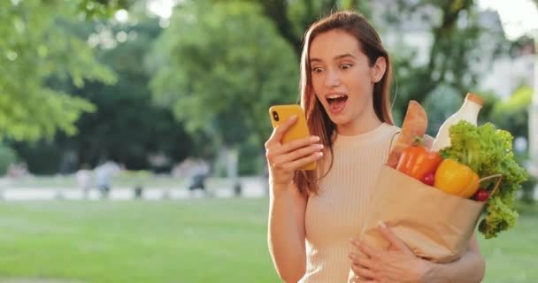 Mujer sorprendida sonriendo mientras está de pie y sosteniendo la bolsa con comida y teléfono. Chica haciendo cara sorprendida mientras mira la pantalla de su teléfono inteligente. Parque borroso y luz solar al fondo . — Vídeos de Stock