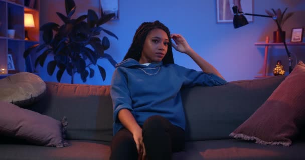 Portret van een jonge Afro-Amerikaanse vrouw die poseert terwijl ze op de bank zit. Aantrekkelijk millennial meisje met dreadlocks draaien hoofd en op zoek naar camera. Gezellige thuis achtergrond. — Stockvideo