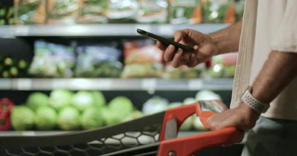 Pemandangan dari tangan manusia memegang smartphone dan layar srolling di supermarket. Guy menggunakan telepon sambil berjalan dan mendorong keranjang belanja dekat rak sayuran. Latar belakang kabur. — Stok Video
