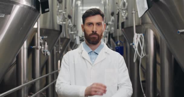 Vista de la cosecha de atento joven trabajador masculino mirando a la cámara Hombre guapo en bata de laboratorio blanco de pie en filas de cubas de cerveza de acero y cruzando las manos.Concepto de fábrica de cerveza y planta de cerveza . — Vídeos de Stock