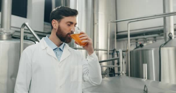 Pohledný pracovník pivovaru, který pije čerstvě uvařené pivo ve skle a jásá. Pohledný muž v bílém plášti zkouší nápoj a dívá se do kamery, zatímco stojí v moderním pivovaru. — Stock video