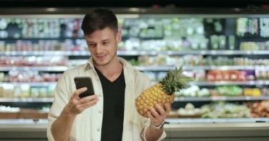 Milenyum adamı ananas ve akıllı telefon ekranına bakarken süpermarkette başını sallıyor ve gülümsüyor. Meyve tutan ve alışveriş yaparken telefon kullanan adam. Bulanık arkaplan.