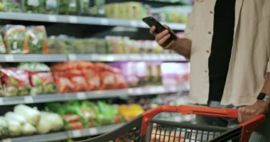 Adamın elinde telefon olan, süpermarkette daktilo eden ve ekrana dokunan görüntüsü. Adam yürürken akıllı telefon kullanıyor ve alışveriş arabasını yeşil market raflarının yakınına itiyor. Bulanık arkaplan.