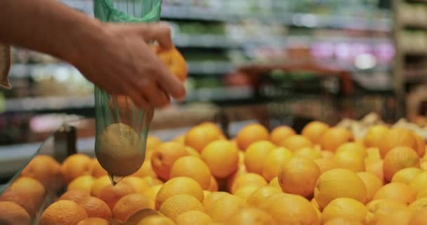 Vue rapprochée de la main de l'homme mettant des oranges dans un sac de production réutilisable. Personne faisant du shopping dans le département des fruits et légumes au supermarché.Concept de mode de vie écologique Fond flou . — Video
