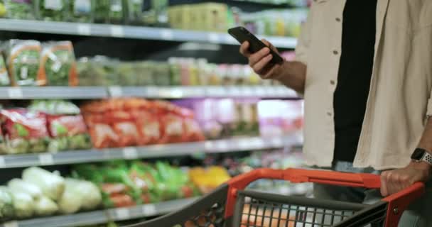 Schnittansicht des Typen mit Telefon, Tippen und Berühren des Bildschirms im Supermarkt. Mann benutzt Smartphone beim Gehen und schiebt Einkaufswagen in der Nähe grüner Lebensmittelregale. Unklarer Hintergrund. — Stockvideo