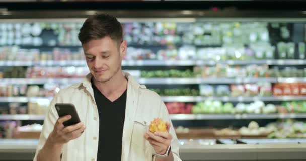 Joven hombre sonriente mirando la pantalla del teléfono inteligente y navegar por Internet mientras está de pie en el supermercado. Millennial hombre sosteniendo kiwano fruta exótica mientras hace compras. Fondo borroso . — Vídeo de stock