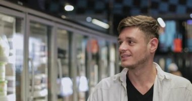 Bin yıllık adamın yürüdüğü ve cam kapılı sergi dolaplarına baktığı yakın plan görüntüler. Genç yakışıklı adam süpermarketin dondurulmuş ürünlerini arıyor. Alışveriş kavramı.