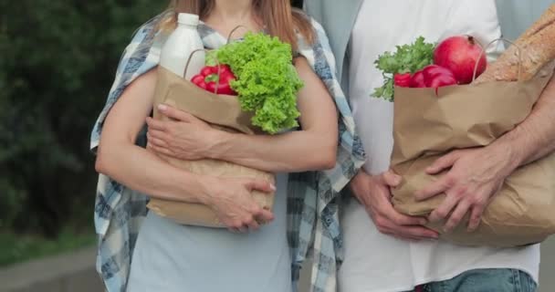 Widok na dziewczynę i faceta trzymających papierowe torby pełne jedzenia i chodzenia po ulicy. Para w luźnych ciuchach, niosąca zakupy w rękach. Pojęcie zdrowego stylu życia. — Wideo stockowe