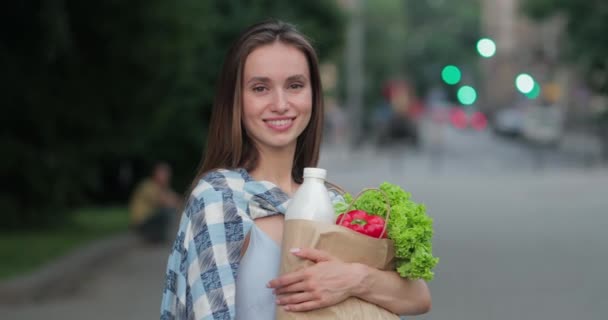 Chica sosteniendo bolsa de papel artesanal con comida y posando mientras está de pie en la calle. Vista de la cosecha de feliz hermosa mujer sonriendo y mirando a la cámara. Concepto de dieta saludable y estilo de vida . — Vídeos de Stock