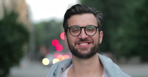 Vista de cerca del hombre guapo en los años 30 mirando a la cámara y sonriendo. Retrato de tipo barbudo alegre en gafas de pie en la calle de la ciudad.Concepto de la vida real y las emociones positivas . — Vídeo de stock