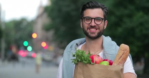 Portrét atraktivního dospělého muže držícího papírový sáček se zdravým jídlem a stojícího na ulici. Oříznout pohled na vousatého chlapíka v neformálních šatech, který vypadá a sedí před kamerou. Venku. — Stock video