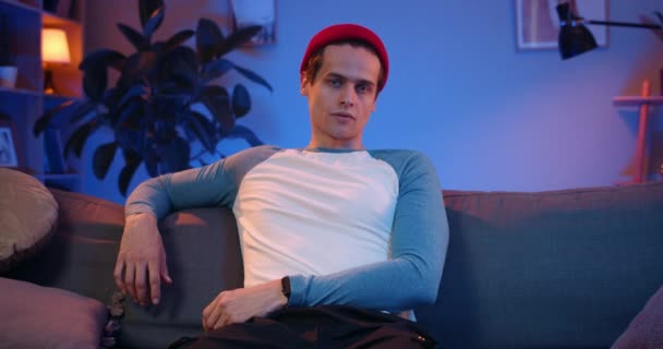 Porträt eines gutaussehenden, ernsthaften jungen Mannes, der auf dem Sofa sitzend in die Kamera blickt. Vorderansicht eines männlichen Millennials mit Hut, der posiert, während er Zeit zu Hause verbringt. Konzept des Lebensstils. — Stockvideo
