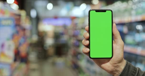 Чоловіча рука показує сучасний чорний смартфон з зеленим екраном у вертикальному положенні на розмитому фоні супермаркету. Концепція ключа хромосоми та сміху . — стокове відео