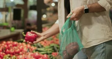 Kırmızı biberi seçen ve tekrar kullanılabilir ürün torbasına koyan insan görüntüsü. Beyaz erkek süpermarketteki meyve ve sebze reyonundan alışveriş yapıyor. Bulanık arkaplan.